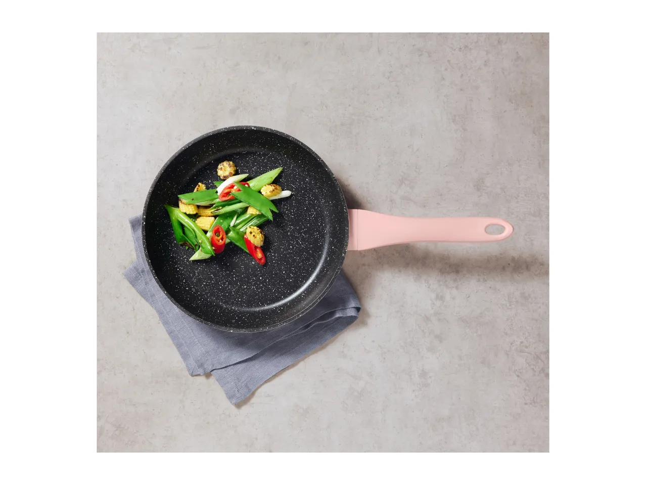 Black frying pan with a pink handle, filled with stir-fried vegetables, on a grey napkin.