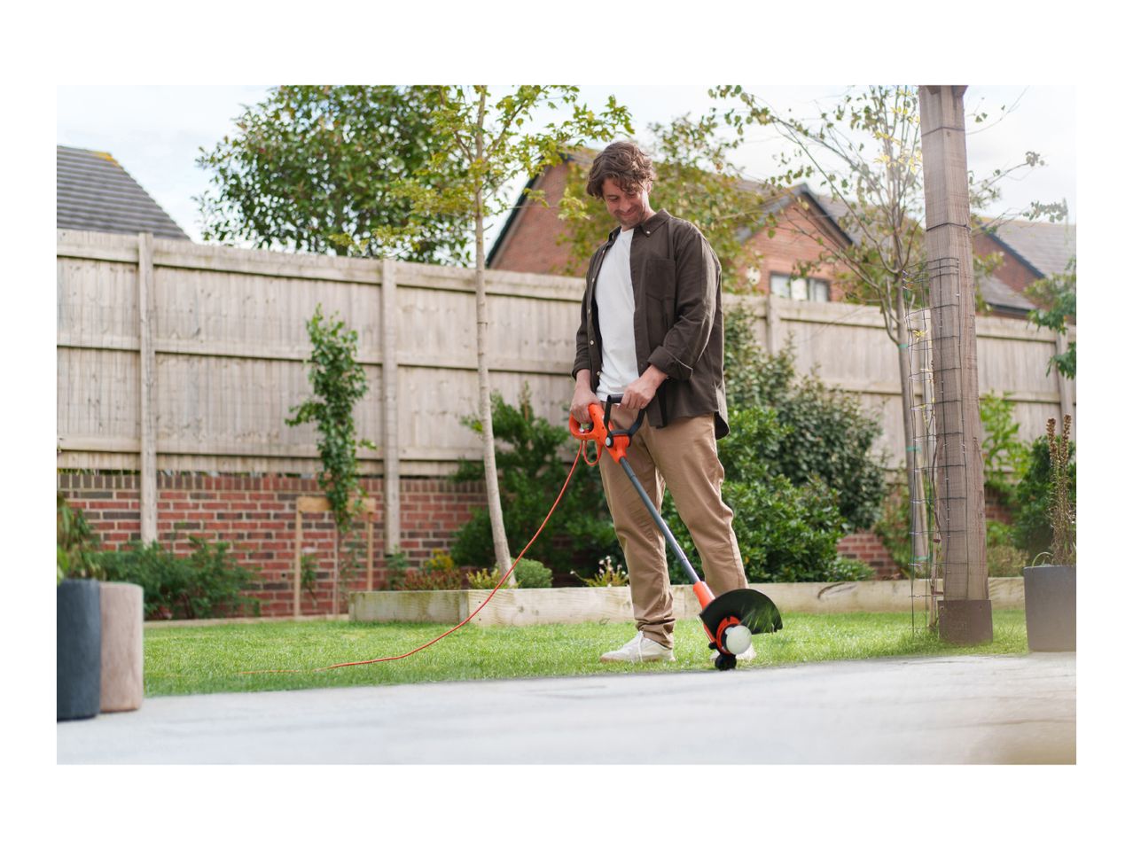 A man using a string trimmer to edge a lawn next to a patio in a backyard.