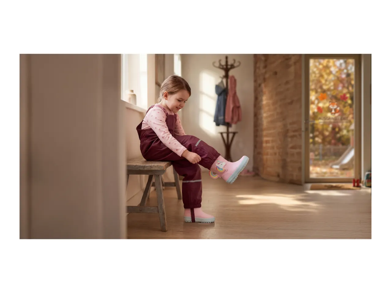 Child in waterproof dungarees and unicorn wellington boots, sitting on a bench indoors.