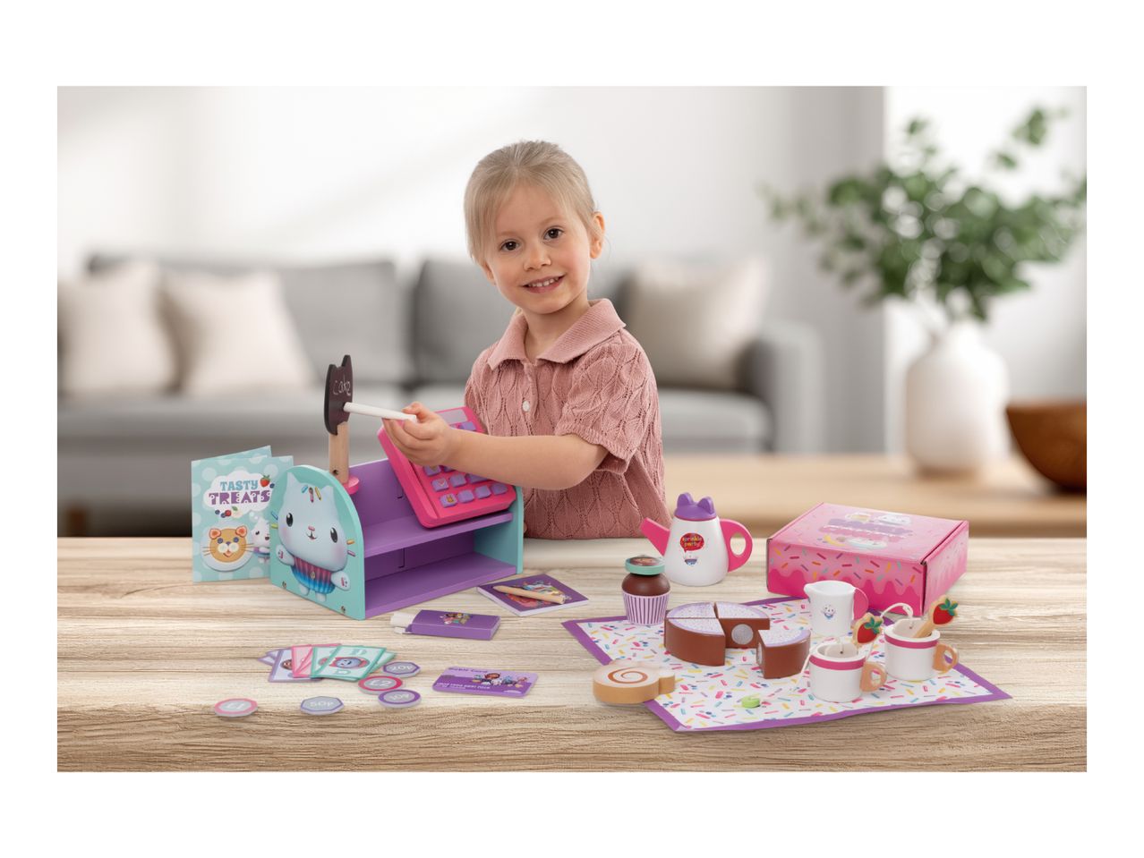 A young girl playing with a toy bakery set, featuring a cash register, cakes, and tea set.