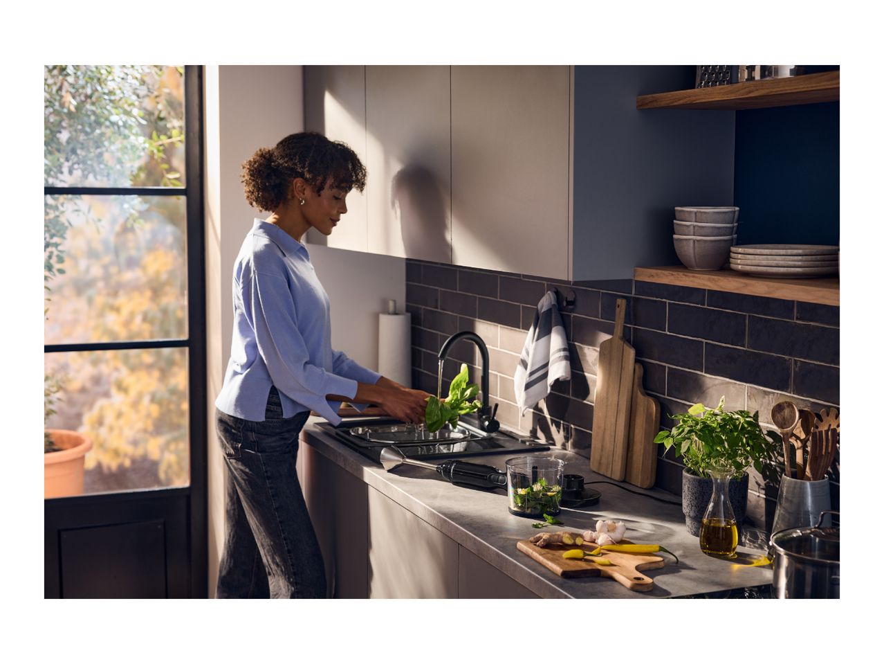 A woman washing fresh herbs at a kitchen sink next to a hand blender and ingredients.