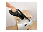 A person uses a black handheld vacuum cleaner to clean crumbs from an open drawer of a wooden nightstand.