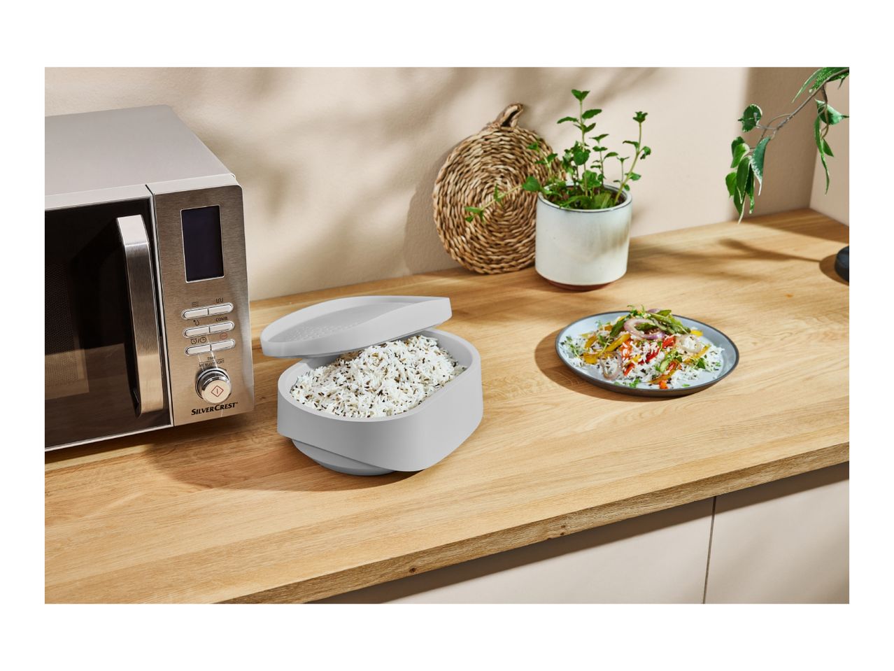 A SilverCrest microwave, a rice cooker, and a plate of rice with vegetables on a wooden kitchen counter.