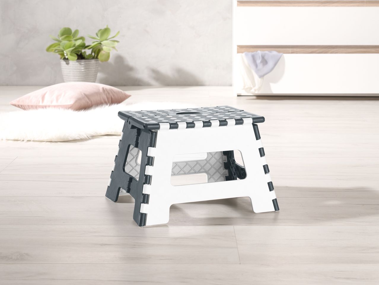 A white and gray plastic folding step stool sits on a light wood floor.