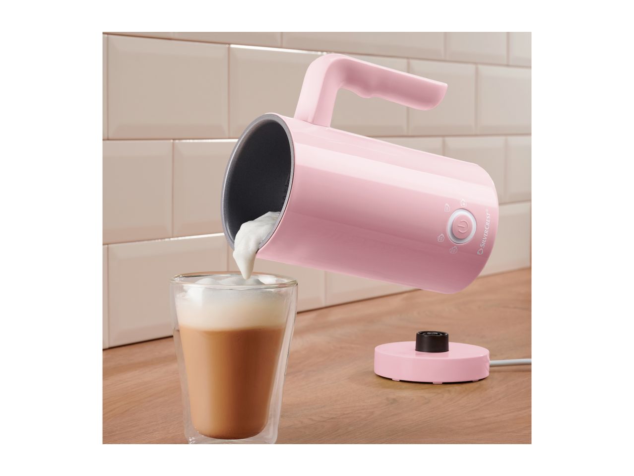Pink milk frother pouring foam into a glass of coffee on a wooden counter.