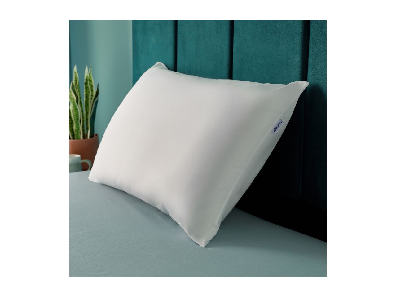 White pillow with a purple tag on a bed with a teal headboard and a plant.