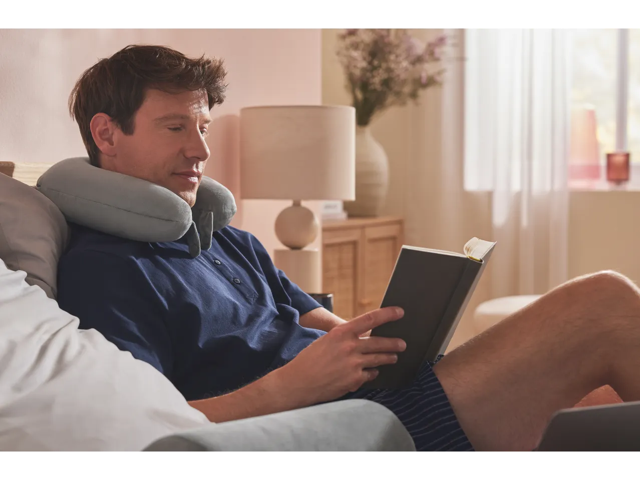 Um homem relaxa na cama com um travesseiro de pescoço, lendo um livro.