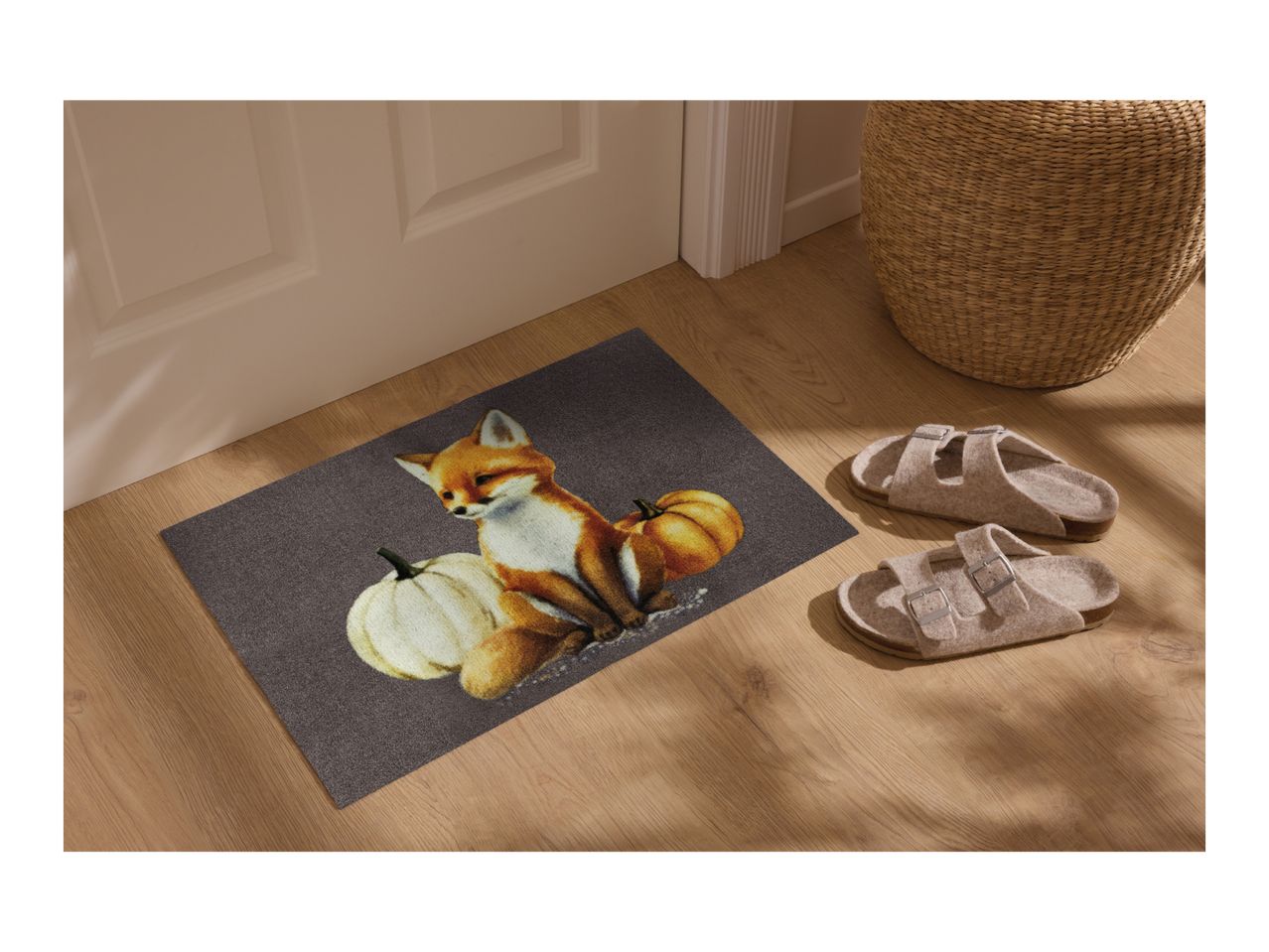 Doormat with a fox and pumpkins, next to a pair of slippers and a woven basket.