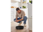 Homem controlando um aspirador robô Silvercrest com um smartphone em uma cozinha.
