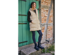 Woman in a beige puffer vest and dark skinny jeans leaning against a green door.