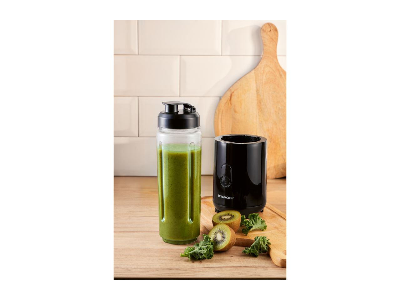 Personal blender with a green smoothie, kiwi, and kale on a wooden board.