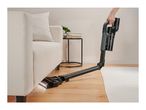 A cordless vacuum cleaner with a flexible wand cleaning under a sofa.