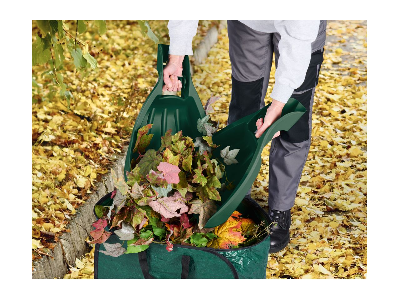 Person using leaf scoops to transfer autumn leaves into a green garden waste bag.