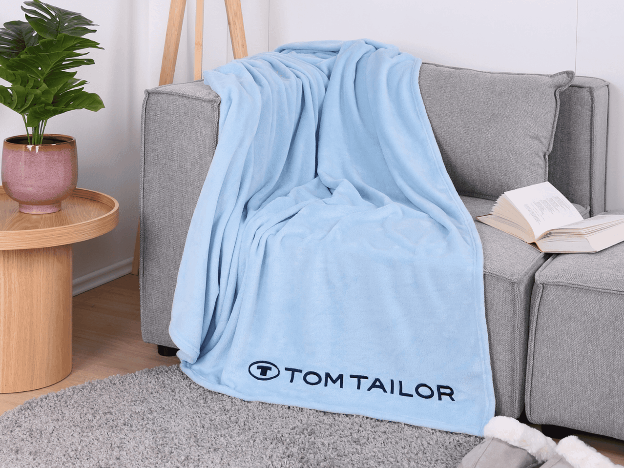 Hellblaue Tom Tailor Fleecedecke auf einem grauen Sofa mit einem Buch und einer Pflanze