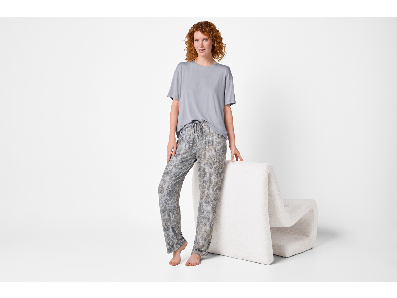 Woman in grey t-shirt and patterned pajama pants, leaning on a white chair.