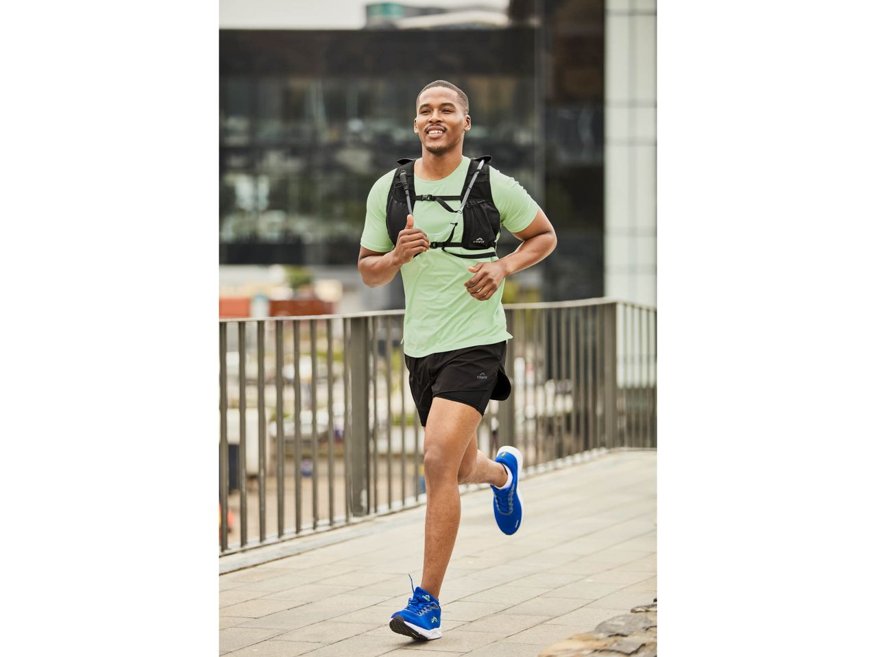 Un uomo sorridente corre indossando una maglietta verde, pantaloncini neri e scarpe da corsa blu.
