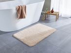 Un tappeto da bagno beige su un pavimento piastrellato grigio accanto a una vasca da bagno bianca.
