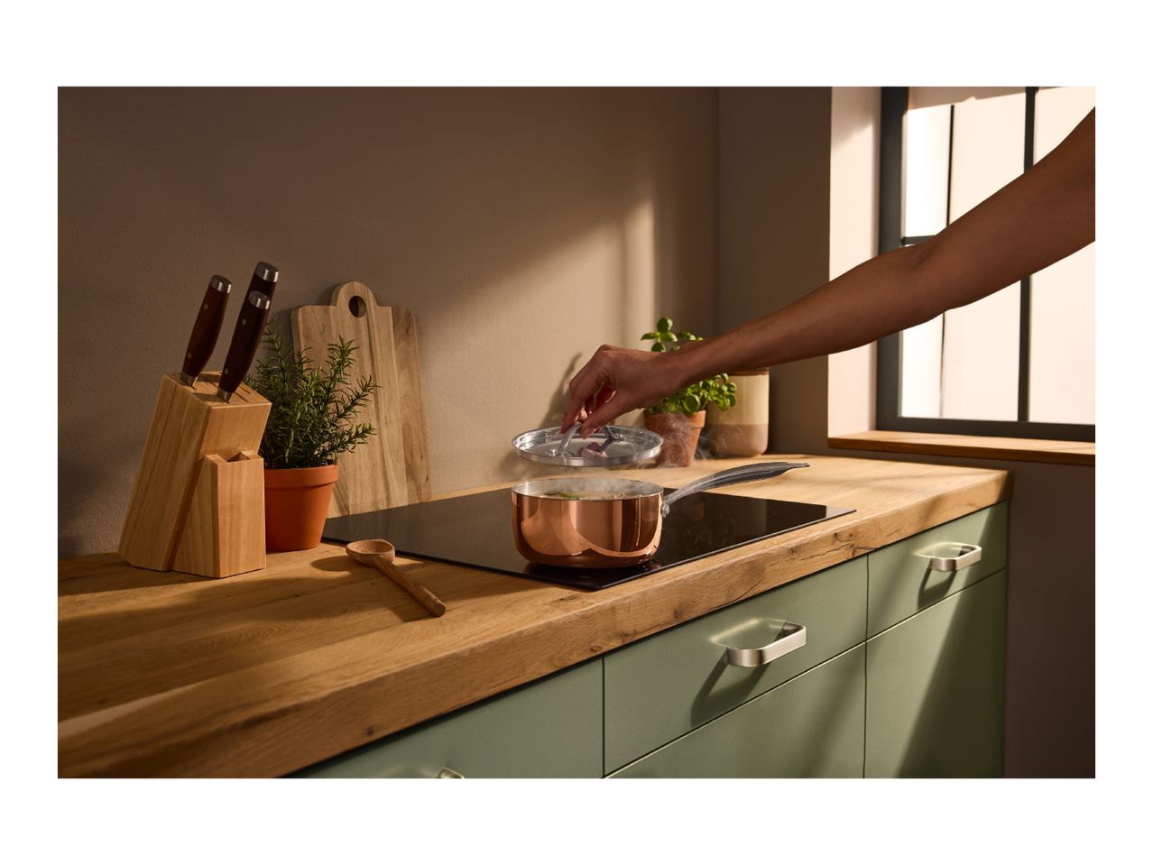 A person lifting the lid of a copper saucepan on an induction hob in a kitchen.