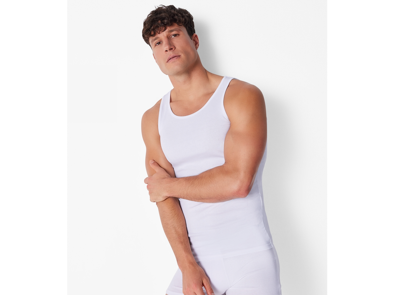 A man in a white ribbed tank top and white briefs, posing against a white wall.