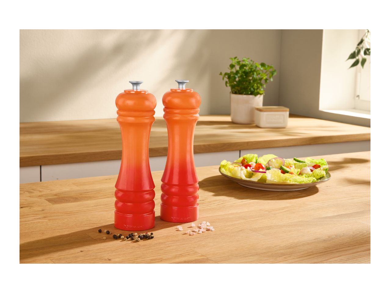 Two orange and red salt and pepper mills on a wooden counter next to a fresh salad.