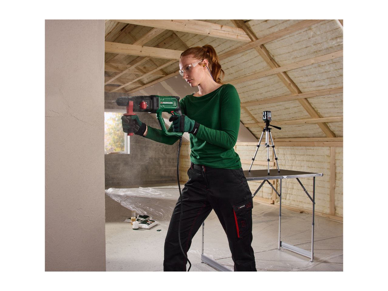 Woman drilling into a wall with a rotary hammer, wearing safety glasses and earplugs, in an attic under construction.