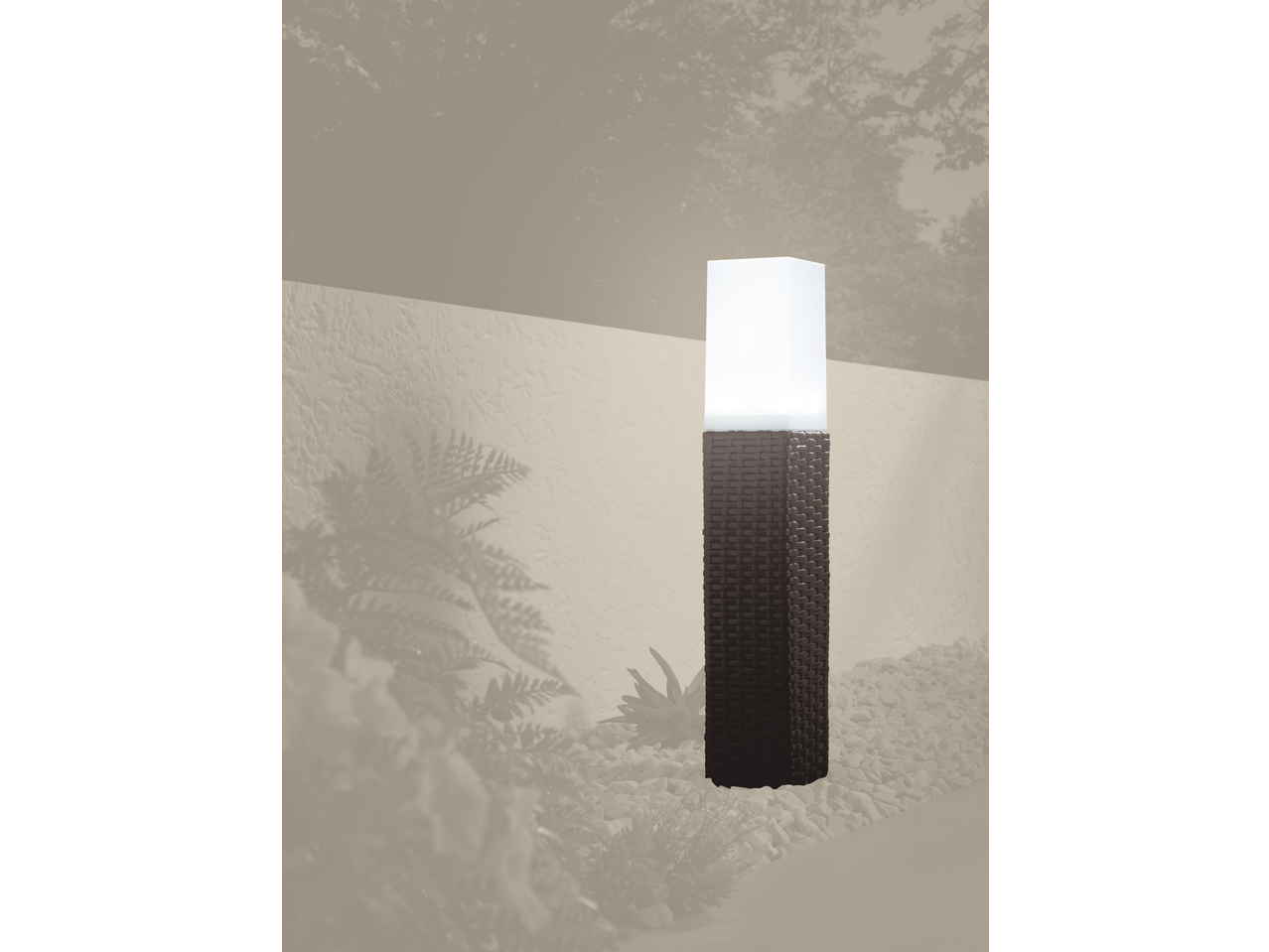 A tall, rectangular outdoor lamp with a dark woven base and a glowing white top, surrounded by white pebbles and plant shadows.