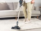 Person vacuuming a light-colored rug with a cordless stick vacuum cleaner.
