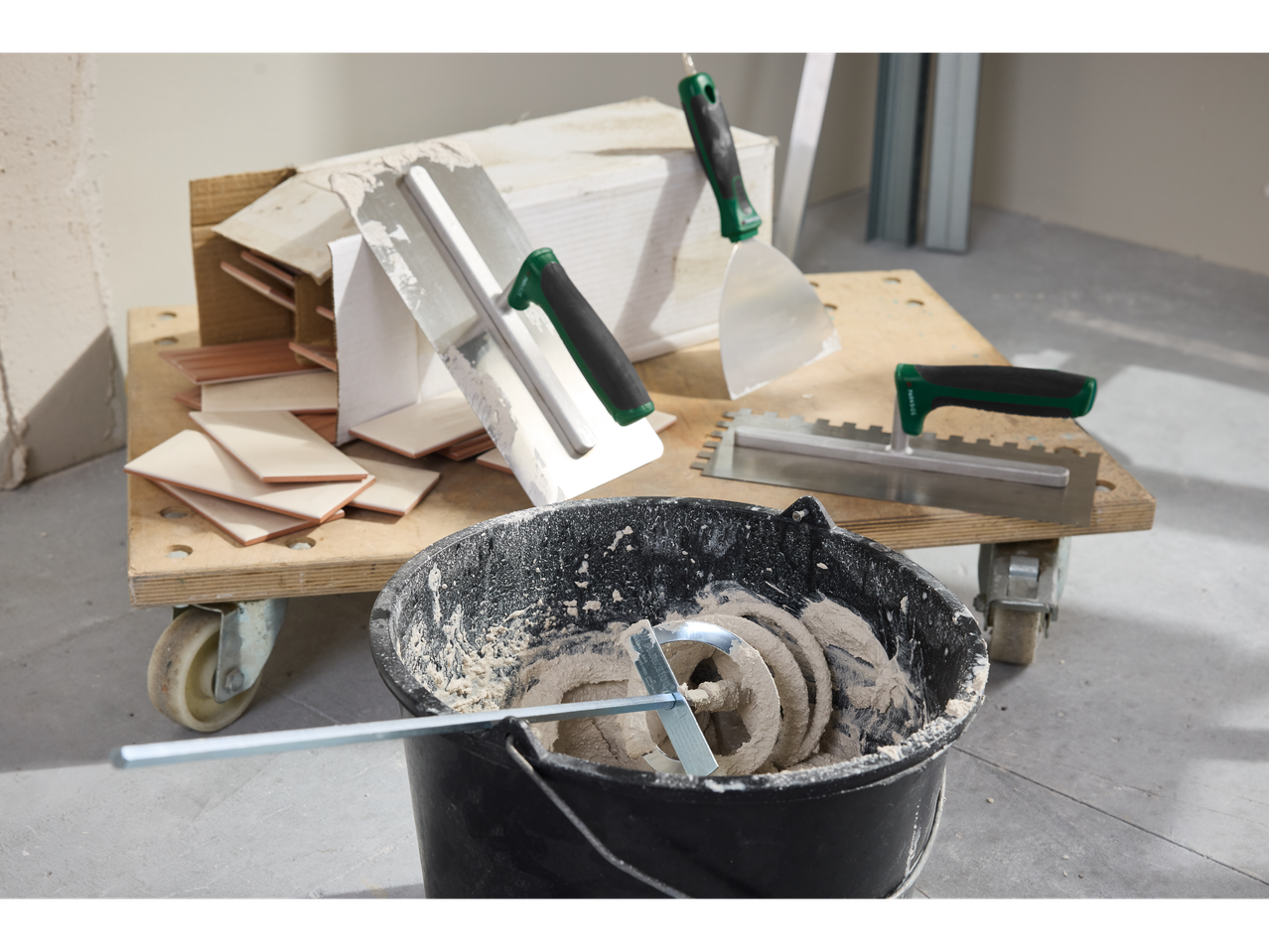 Tiling tools and materials, including trowels, spatulas, and a bucket of mixed mortar, on a wooden cart.
