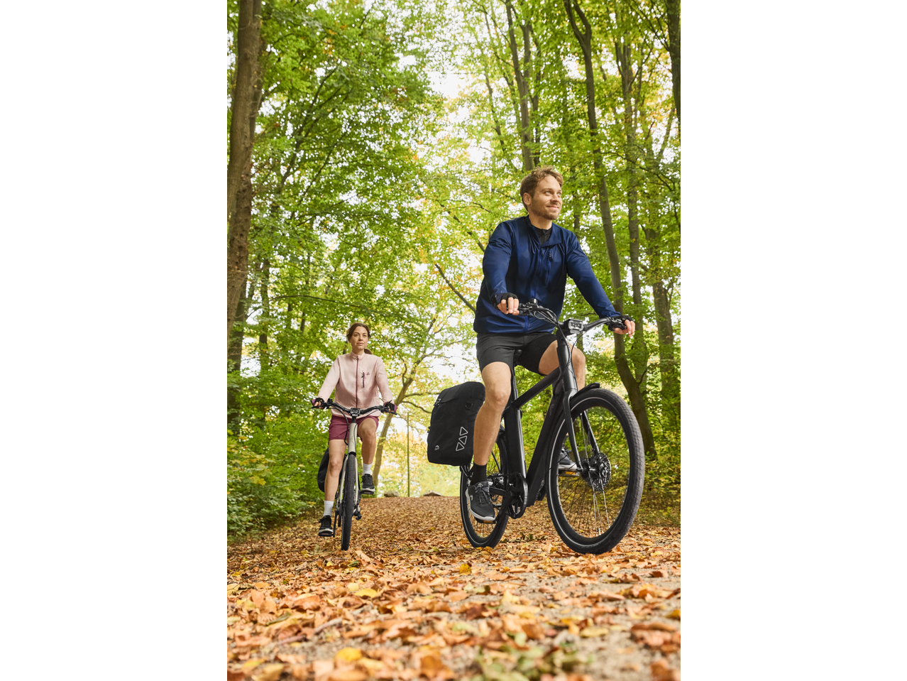 Two people cycling on electric bikes with pannier bags through an autumn forest.