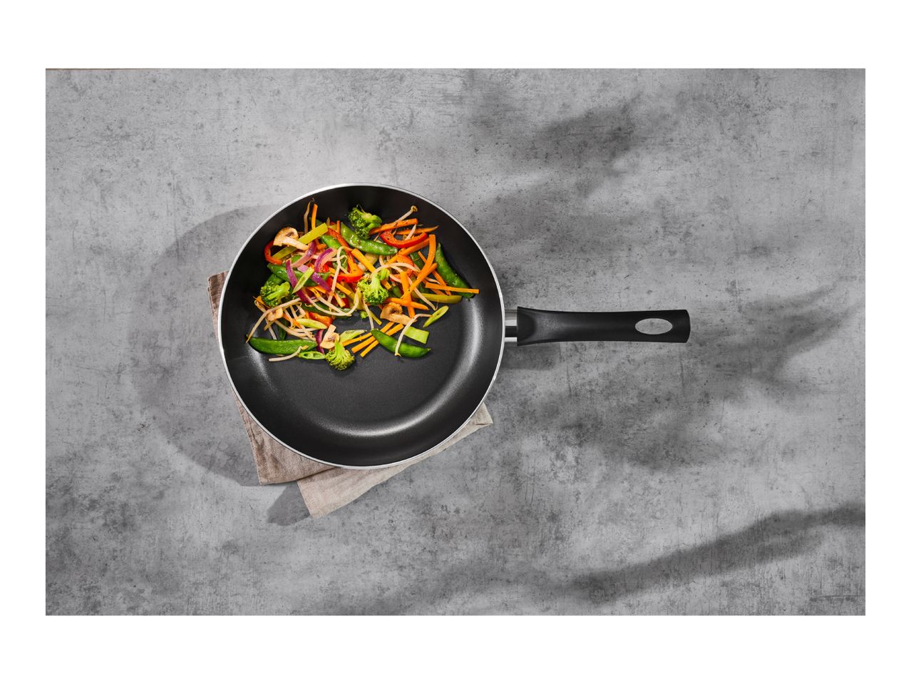 Frying pan with colorful stir-fry vegetables on a grey concrete surface.
