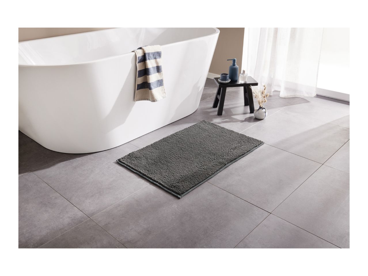 A modern bathroom with a white freestanding bathtub, a gray bath mat, and a small black stool.