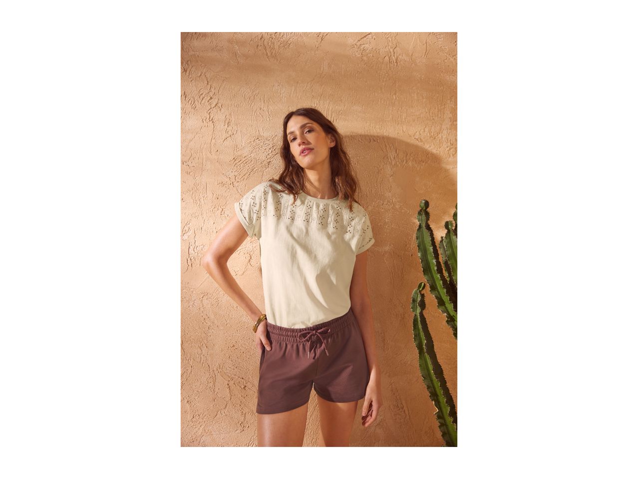 Woman in a cream eyelet t-shirt and brown shorts, standing next to a cactus.