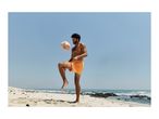 Man in orange swim shorts juggling a soccer ball on a sandy beach with ocean in the background.
