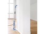 A spray mop, rests against a wall, cleaning a light wood floor.