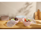 Pink bowls with salad and fruit, with transparent lids, on a wooden table.