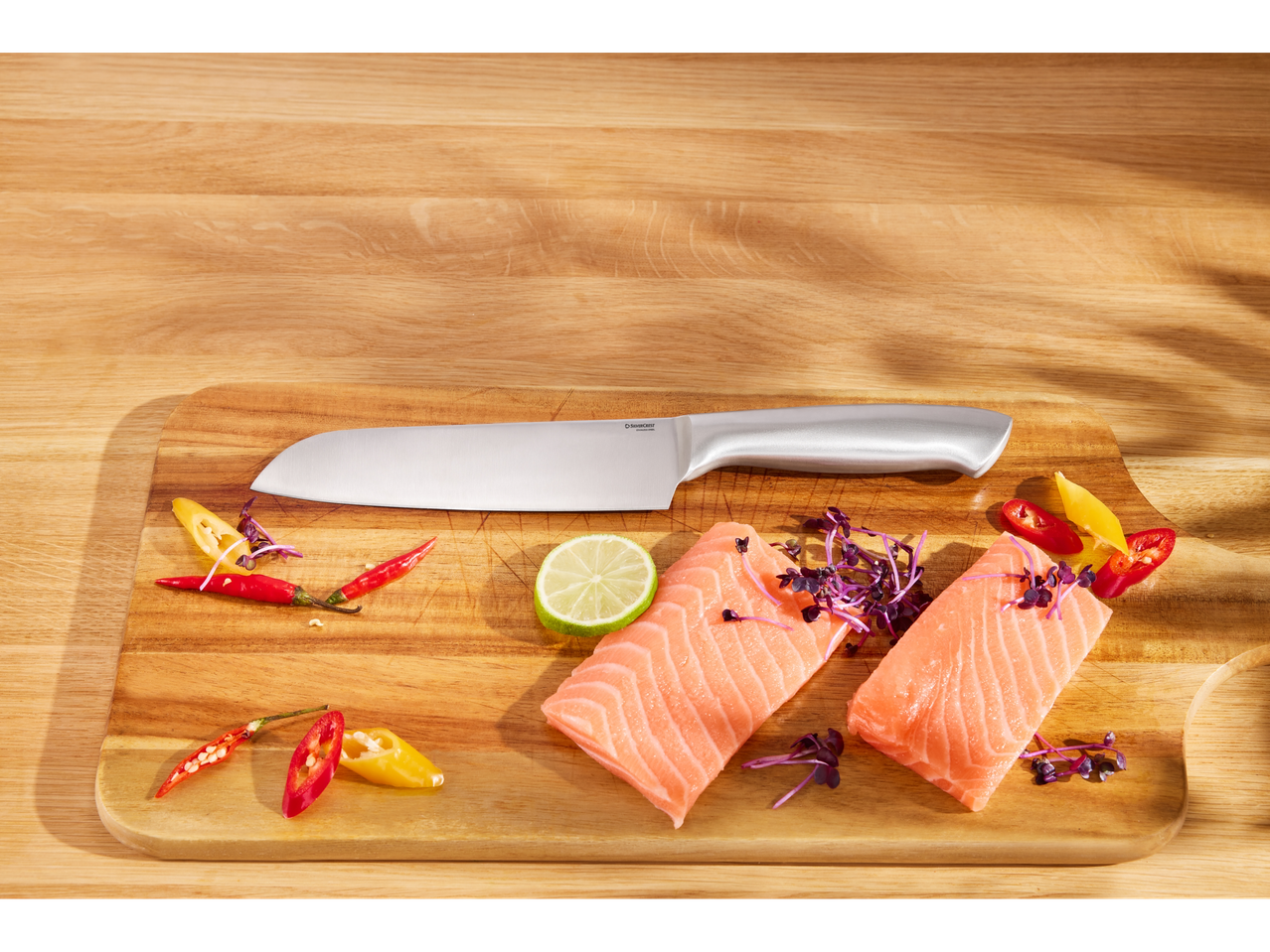 Due filetti di salmone, peperoncini, lime e un coltello Silvercrest su un tagliere di legno.