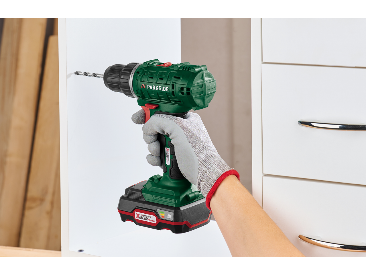 Bormașină verde fără fir cu burghiu, ținută de o mână înmănușată, care găurește un raft alb.