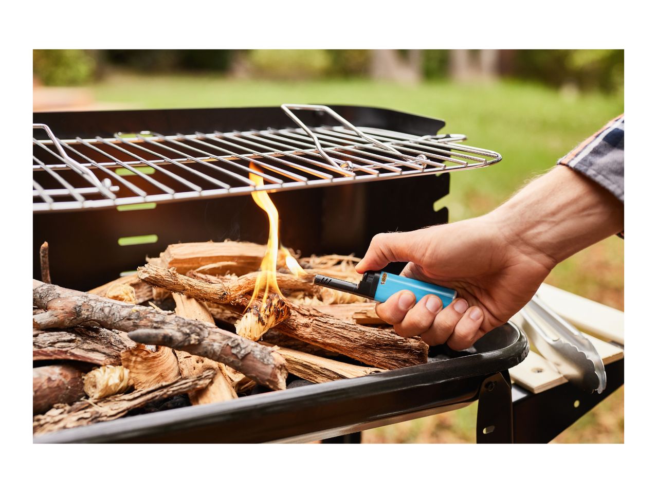 A hand lighting wood in a barbecue grill with a blue utility lighter.