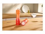 Two orange and red pepper mills on a wooden kitchen counter with a bottle of oil and a towel.