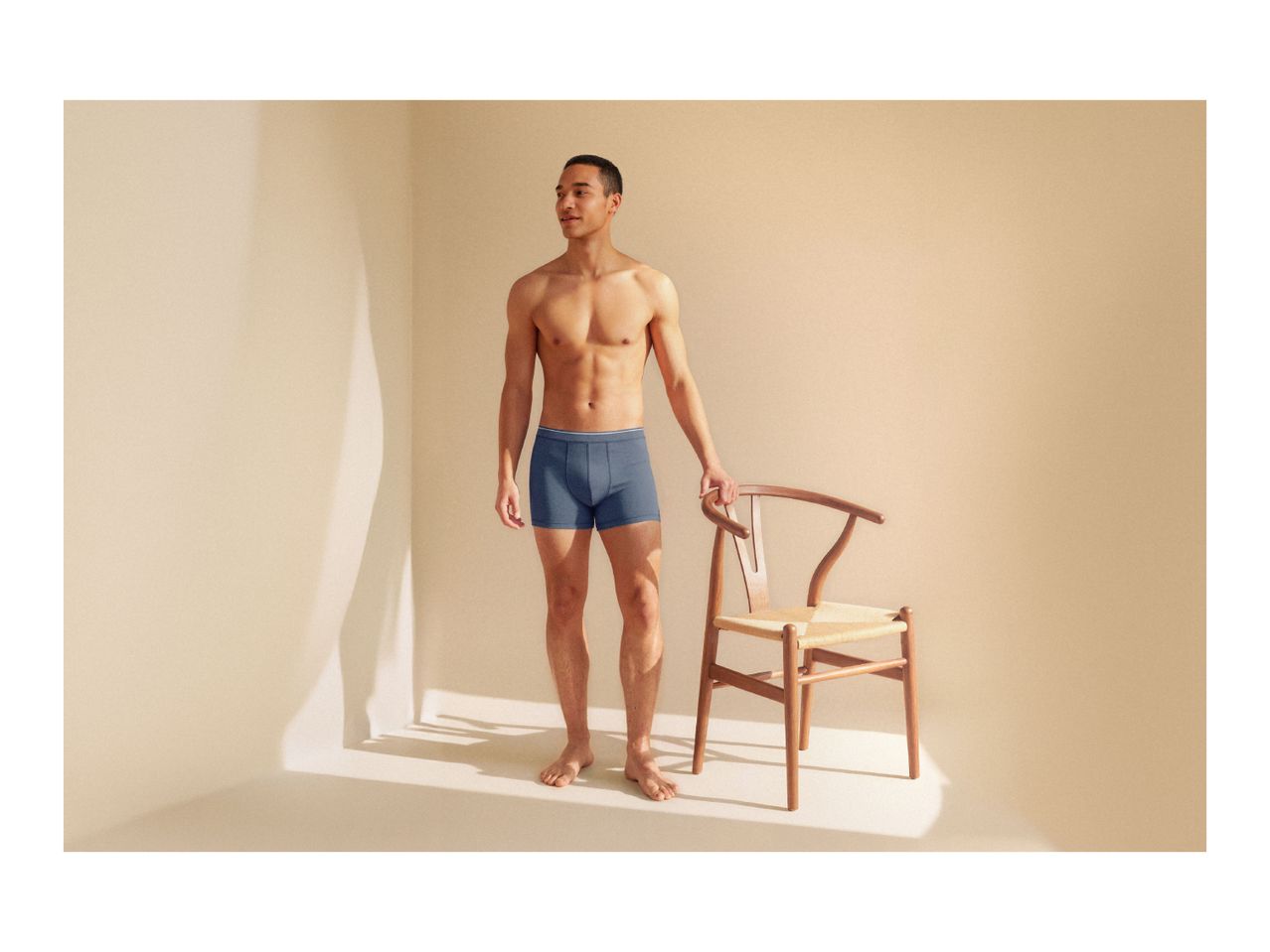 Man in blue boxer briefs standing next to a wooden chair in a sunlit room.