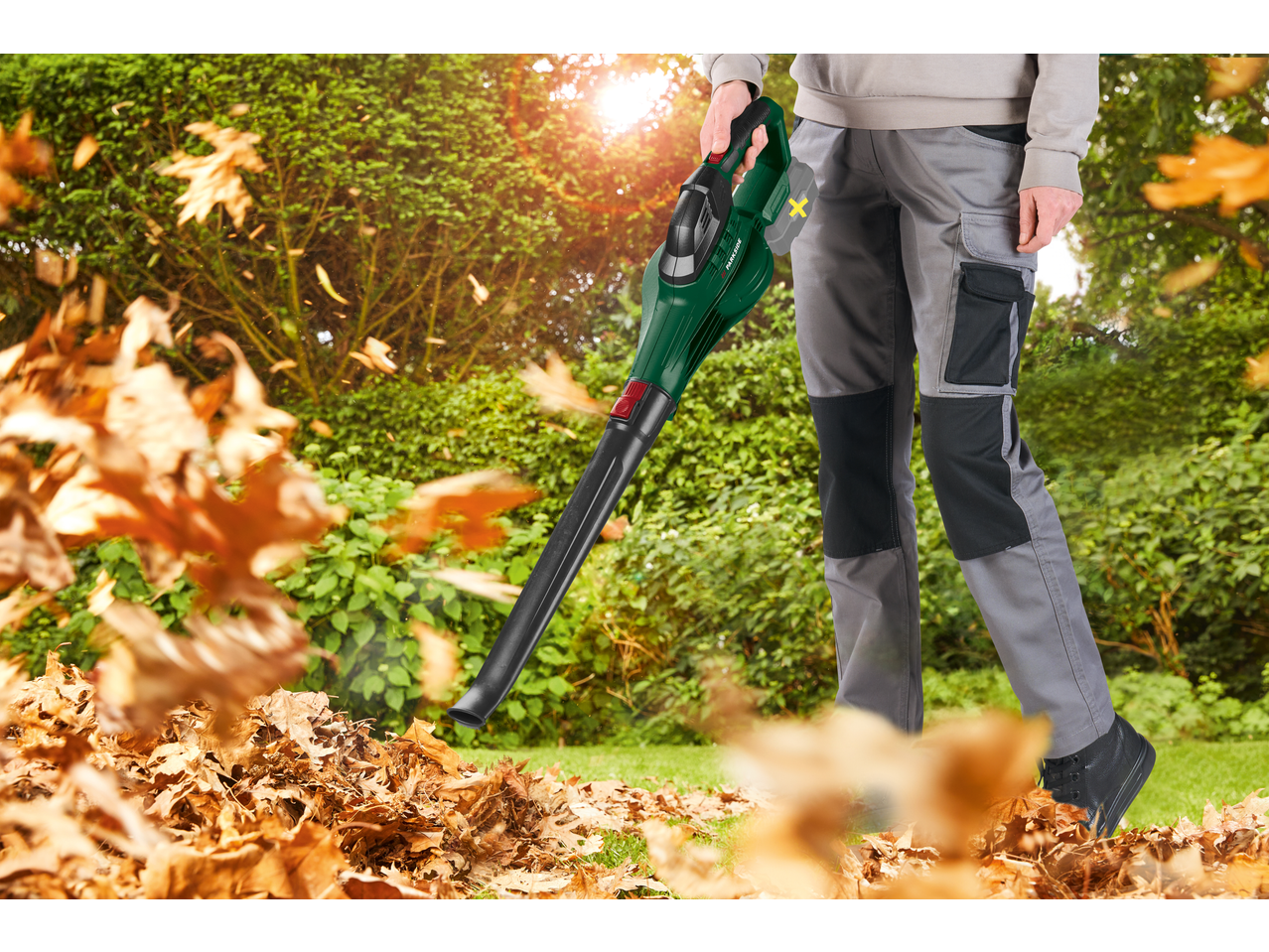 Person using a leaf blower to clear a pile of autumn leaves in a garden.