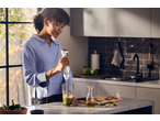 Woman blending green ingredients in a glass with an immersion blender in a modern kitchen.