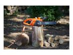 Orange electric chainsaw on a tree stump with cut logs nearby in a garden setting.