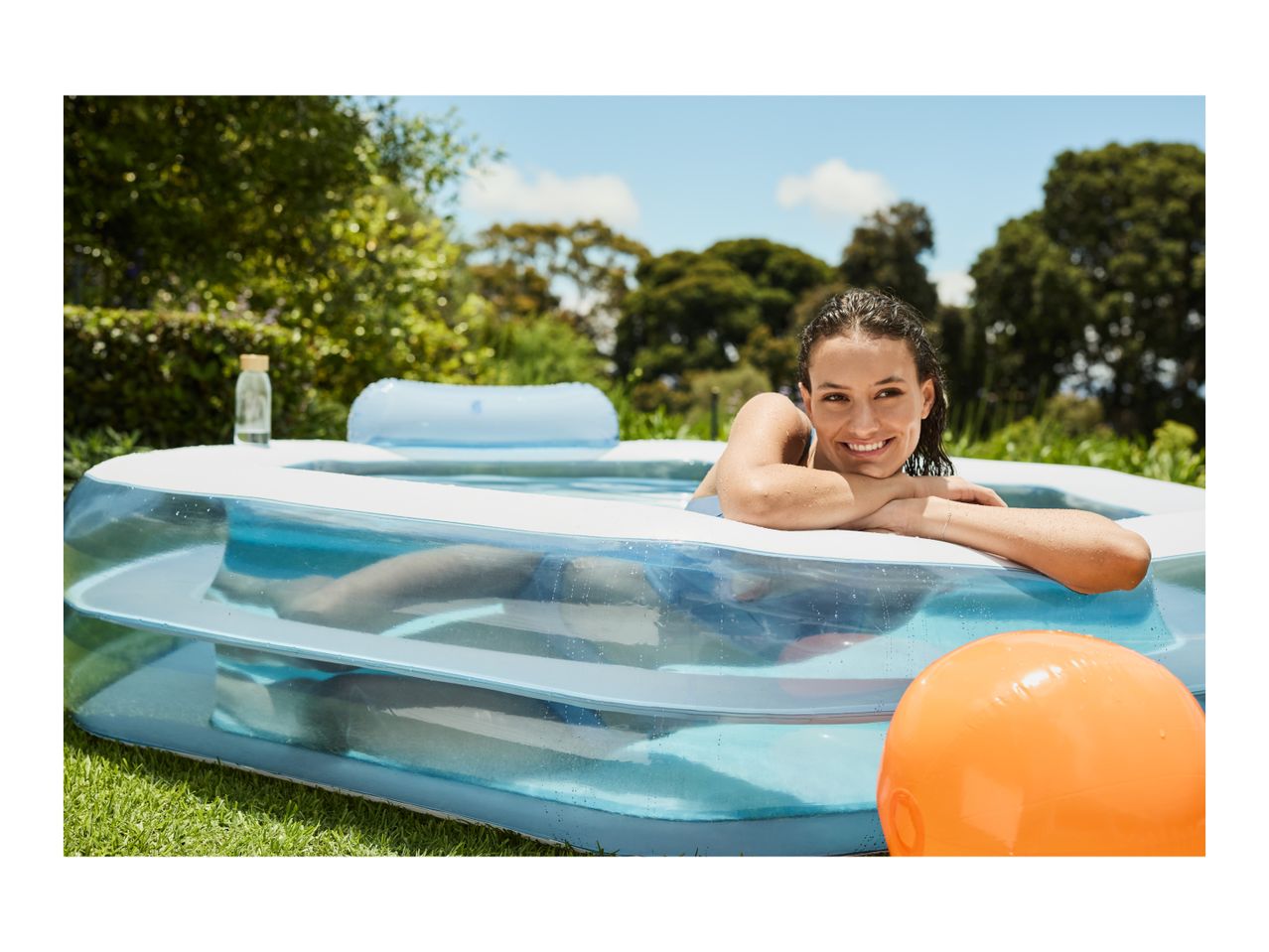 Woman relaxing in an inflatable pool in a garden with an orange ball.