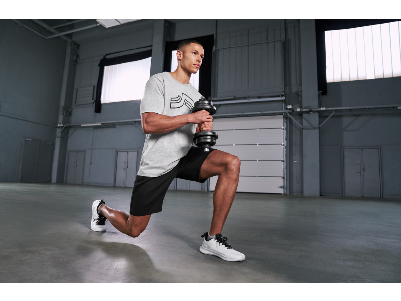 Muškarac u Crivit kratkim hlačama i sivoj majici, radi iskorake s bučicom u teretani.