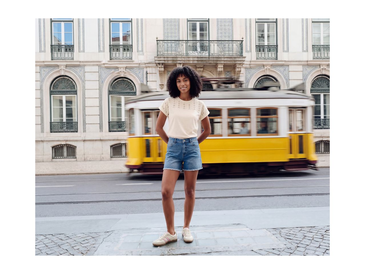 Woman in cream t-shirt and denim shorts, smiling in front of a yellow tram.