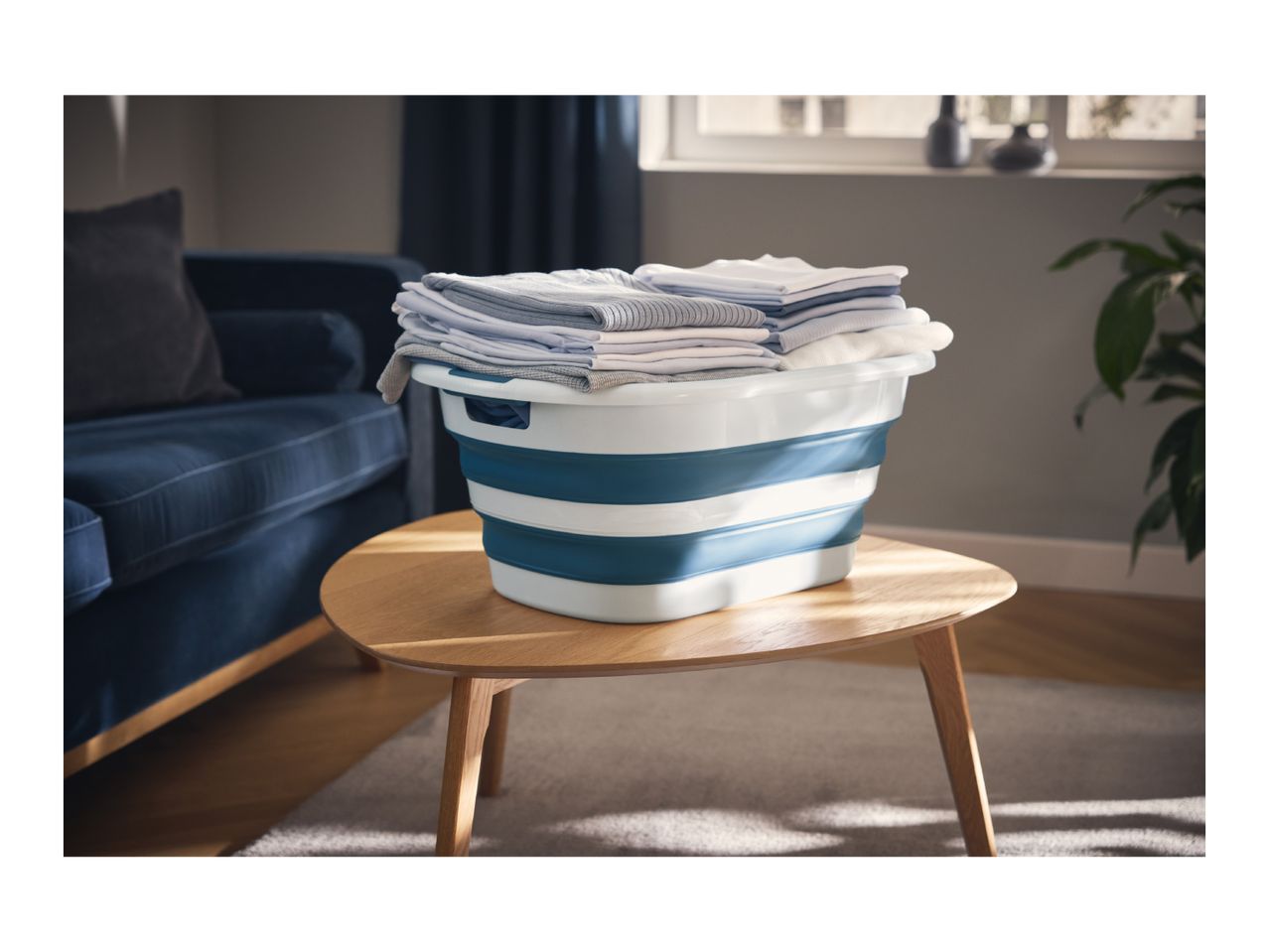 Foldable laundry basket with folded clothes on a wooden table in a living room.