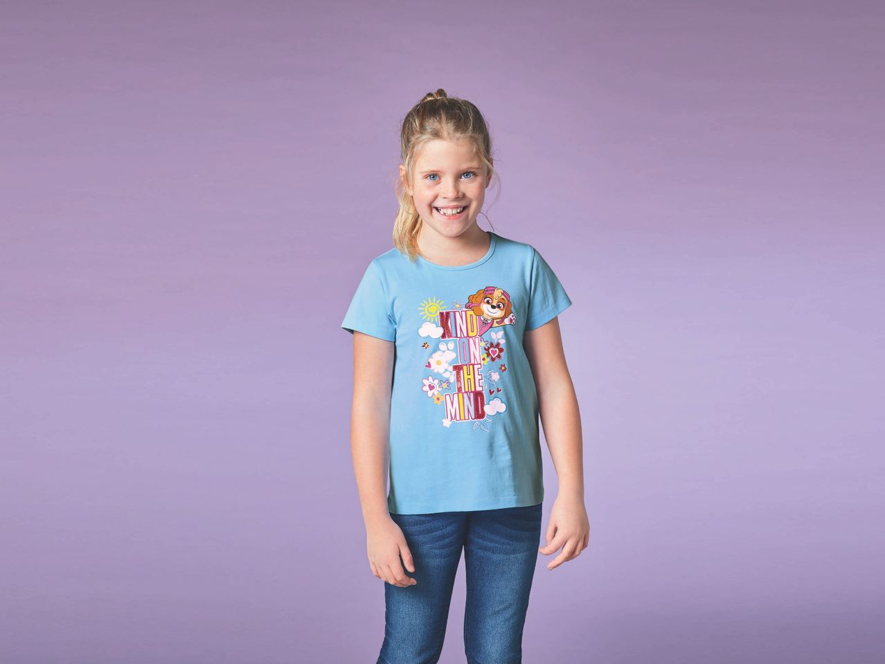Una bambina sorridente indossa una maglietta azzurra con stampa Paw Patrol e jeans.