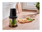 Food processor with basil and oil, next to cheese cubes, pine nuts, and fresh basil on a wooden board.