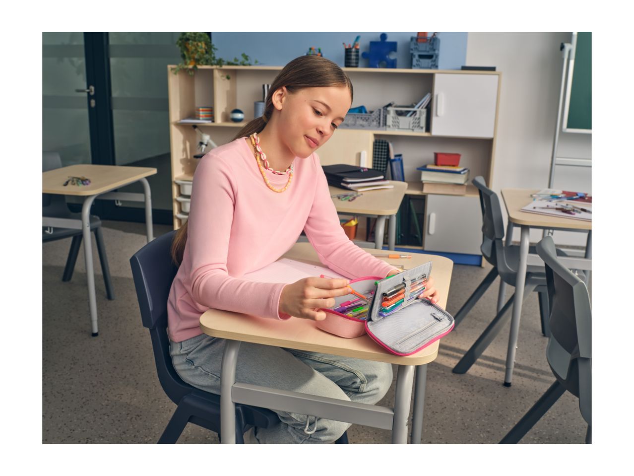 A girl in a classroom, wearing a pink long-sleeve shirt, selecting a pen from a pencil case.